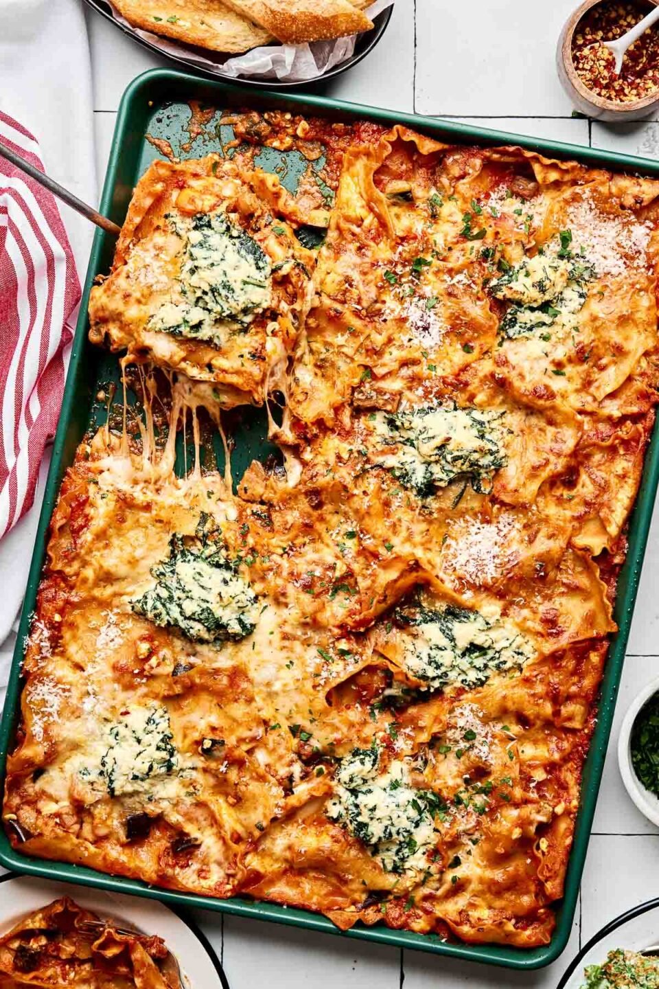 A baking sheet filled with cheesy baked lasagna, cut into squares. Some pieces are being served, showing stretchy melted cheese, tomato sauce, and dollops of creamy spinach ricotta on top. A red and white towel is beside the tray.